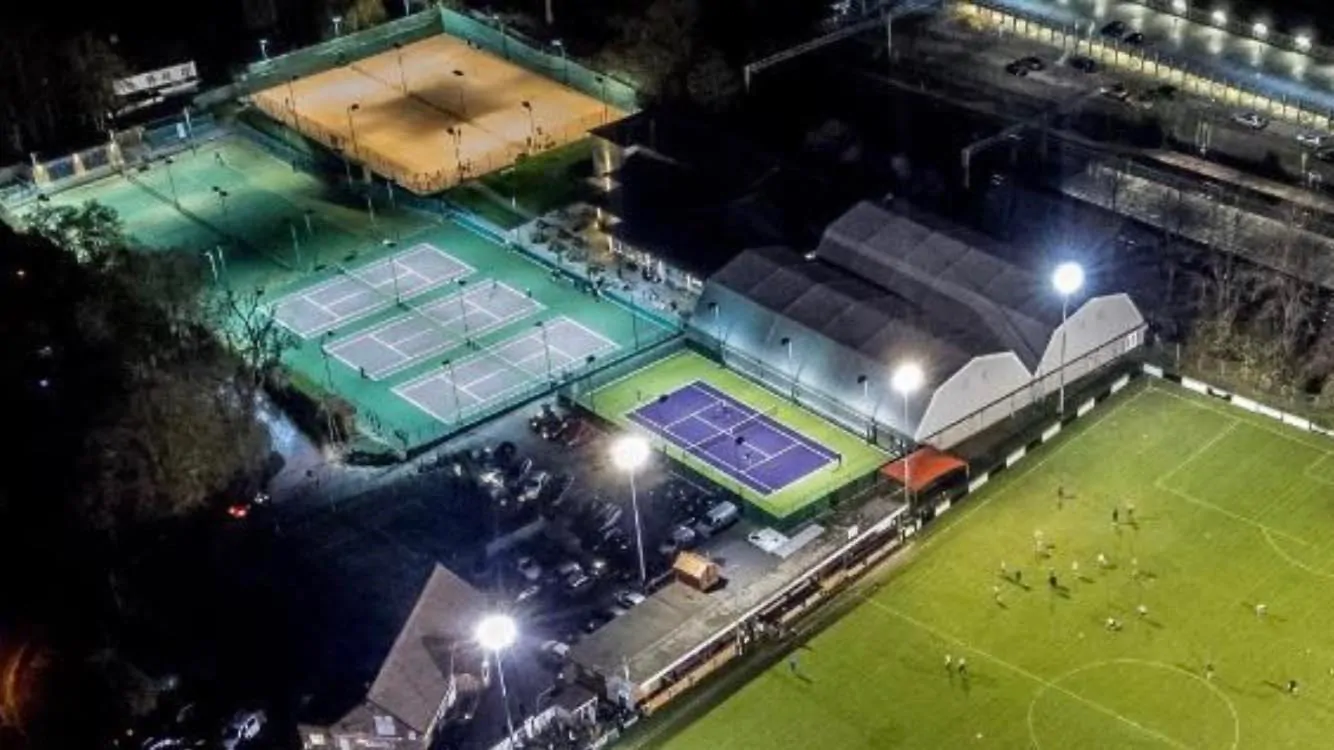 A drone view of the tennis courts and neighbouring football pitch, lit up by floodlights at night.